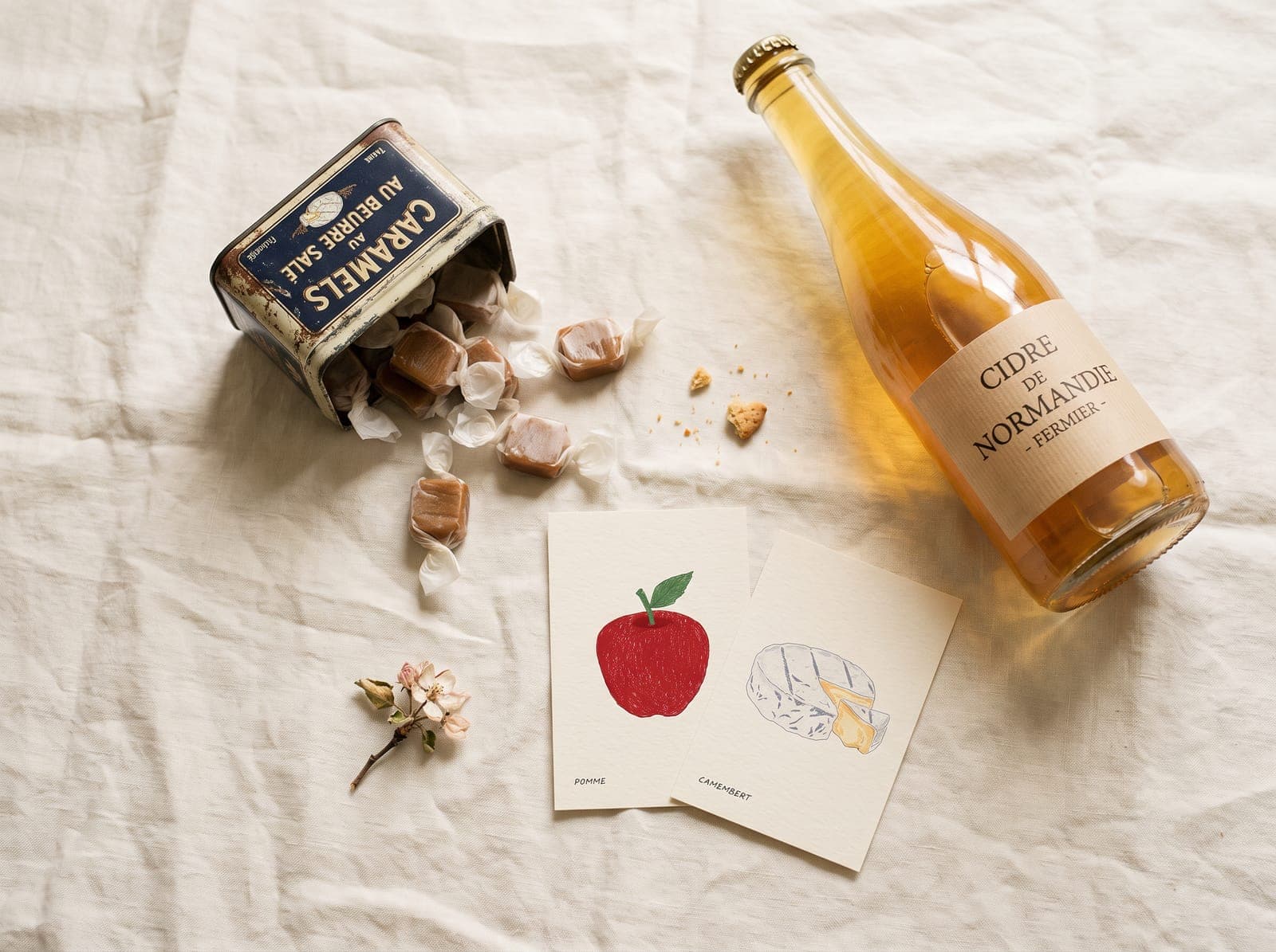 Caramels au beurre salé, bouteille de cidre fermier et deux cartes illustrées Pomme et Camembert sur une nappe en lin